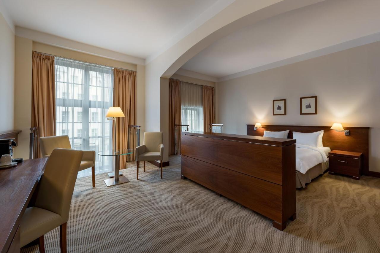 Radisson Blu Hotel, Гданск Екстериор снимка The photo shows a well-furnished hotel room featuring a large bed with a wooden headboard positioned in the center. There are two chairs and a small table on one side, creating a cozy seating area. Near the window, which is covered with light curtain