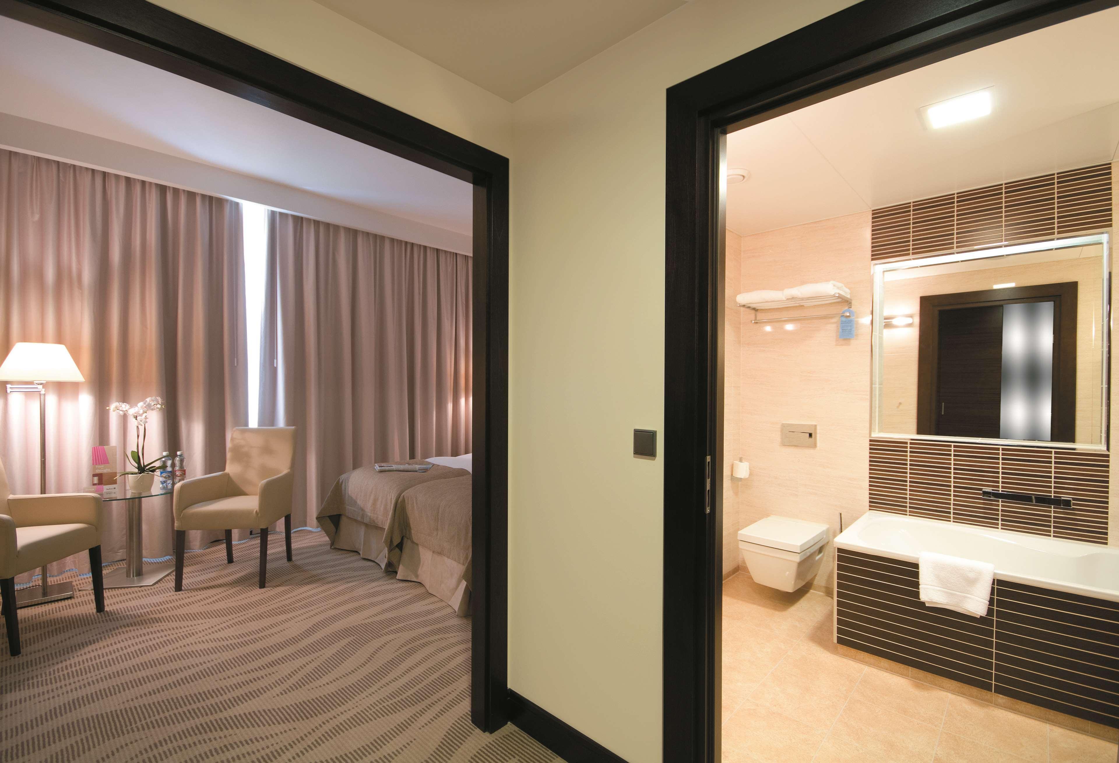 Radisson Blu Hotel, Гданск Екстериор снимка The photo shows a view of a room with two doors leading to different areas. On the left side, there's a bedroom featuring a bed with a light gray coverlet, complemented by curtains and a small seating area with a chair and a table. On the right side,