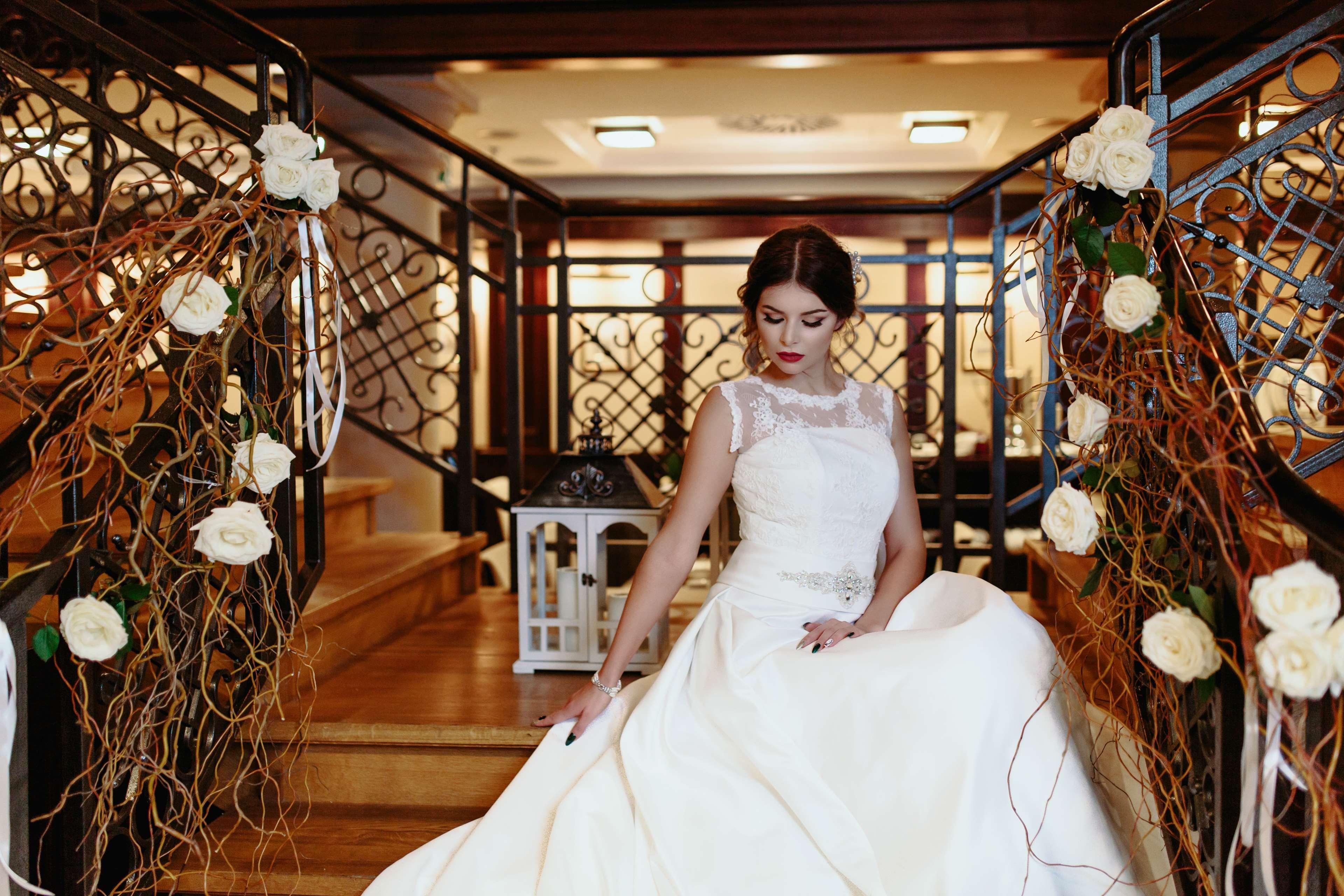 Radisson Blu Hotel, Гданск Екстериор снимка The image shows a bride seated on a staircase in an elegant indoor setting. She is wearing a beautiful white wedding gown with intricate details, including a sheer neckline. The staircase is adorned with decorative elements, such as white roses and f