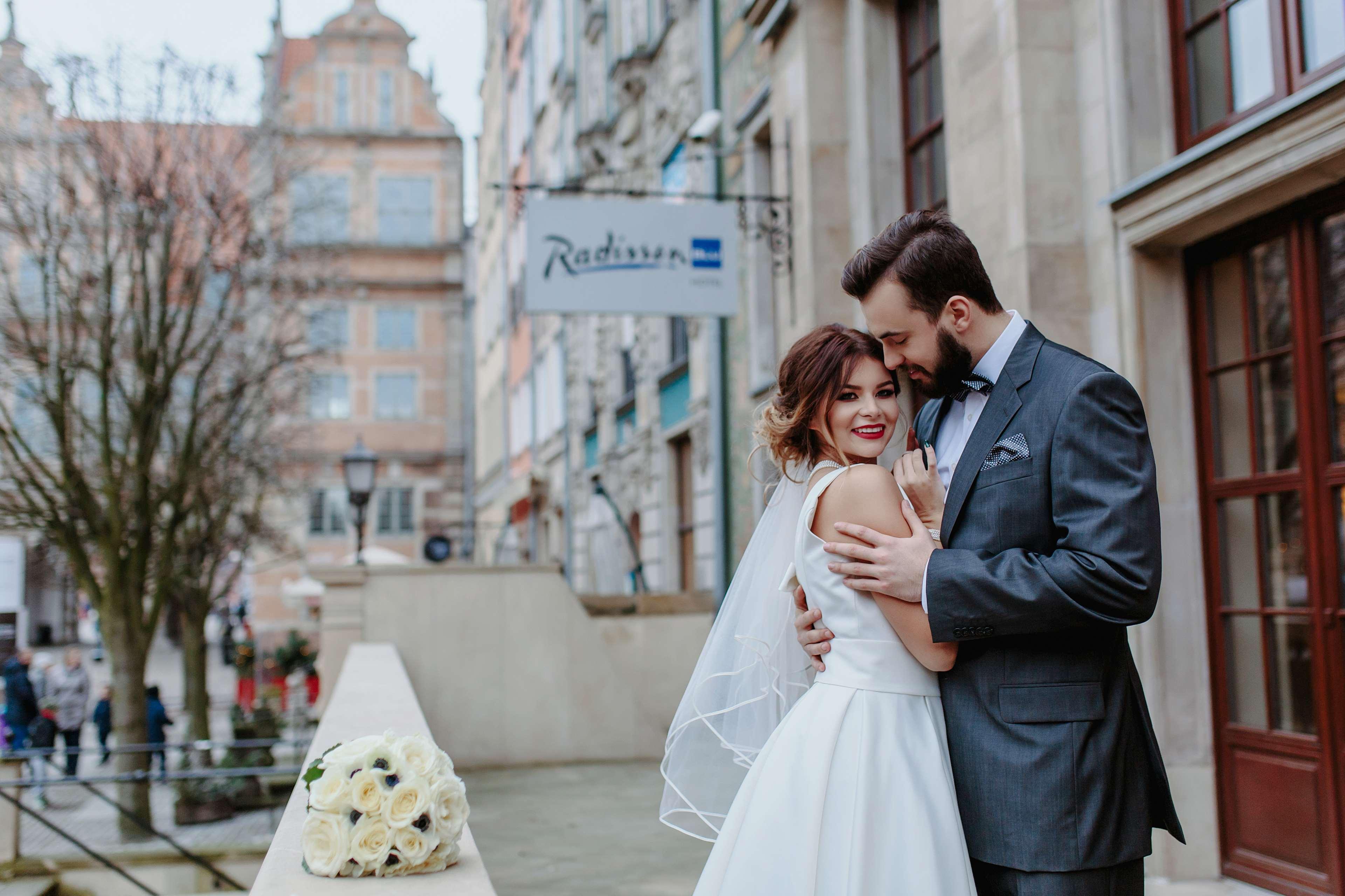 Radisson Blu Hotel, Гданск Екстериор снимка The photo depicts a bride and groom embracing on a balcony. The bride is wearing a white wedding dress with a veil, and she is smiling while leaning into the groom. He is dressed in a grey suit and is holding her close, both appearing very happy. In 