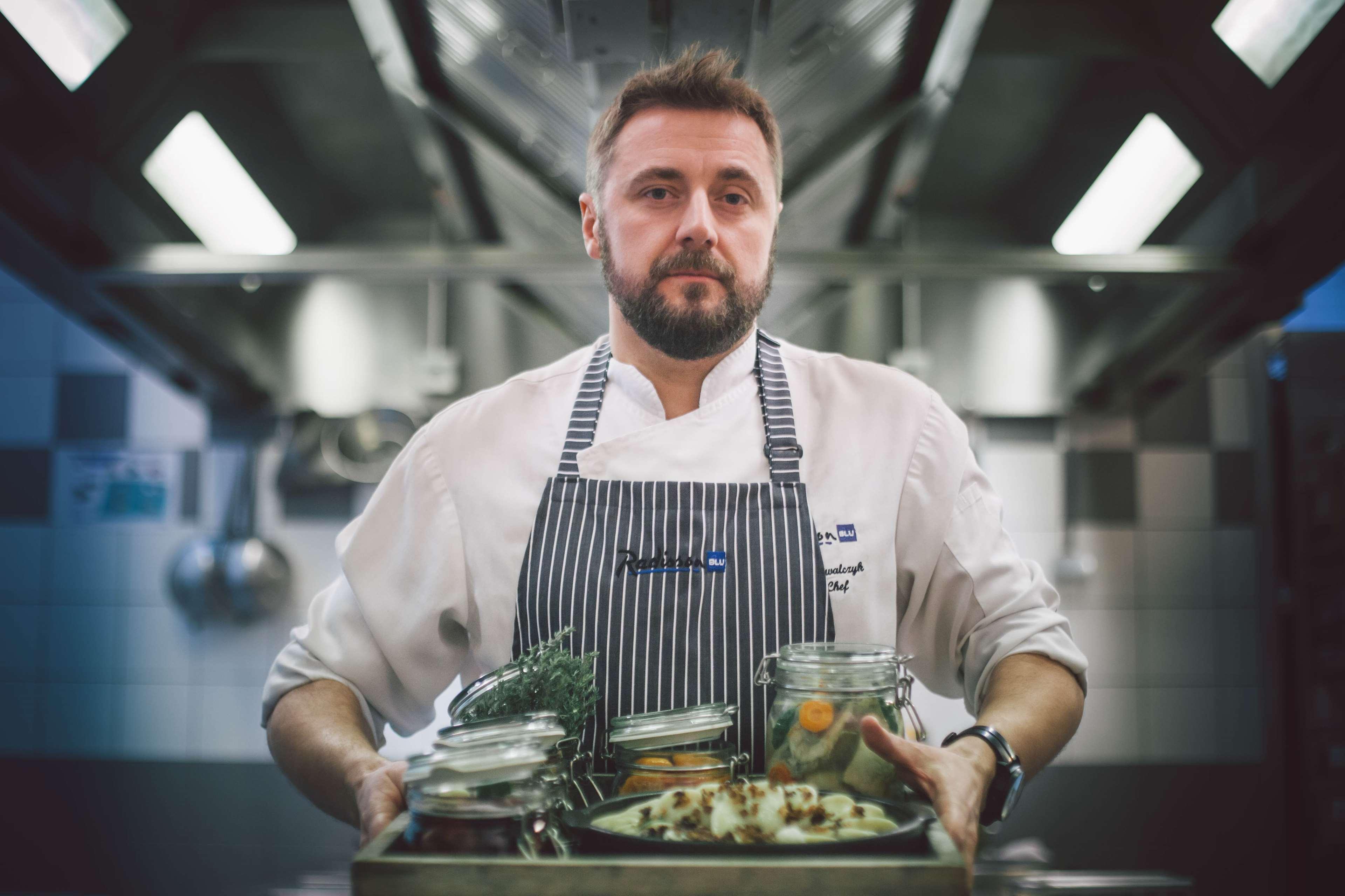 Radisson Blu Hotel, Гданск Екстериор снимка The photo depicts a chef in a professional kitchen setting. He is wearing a white chef's coat with a striped apron and has a focused expression. He is holding a wooden tray filled with various jars and containers that likely contain ingredients for c