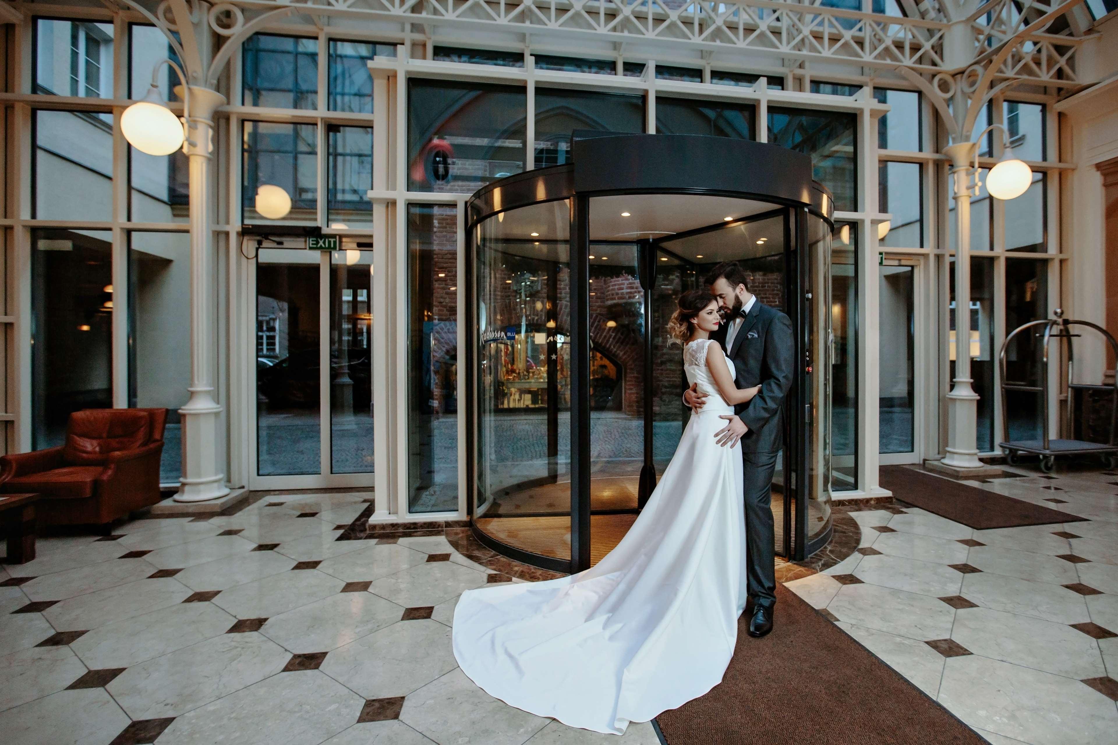 Radisson Blu Hotel, Гданск Екстериор снимка The photo shows a couple in formal attire standing outside a building, likely a hotel or venue. They appear to be newlyweds, with the woman in a beautiful white wedding gown and the man in a suit. They embrace each other tenderly in front of a glass 