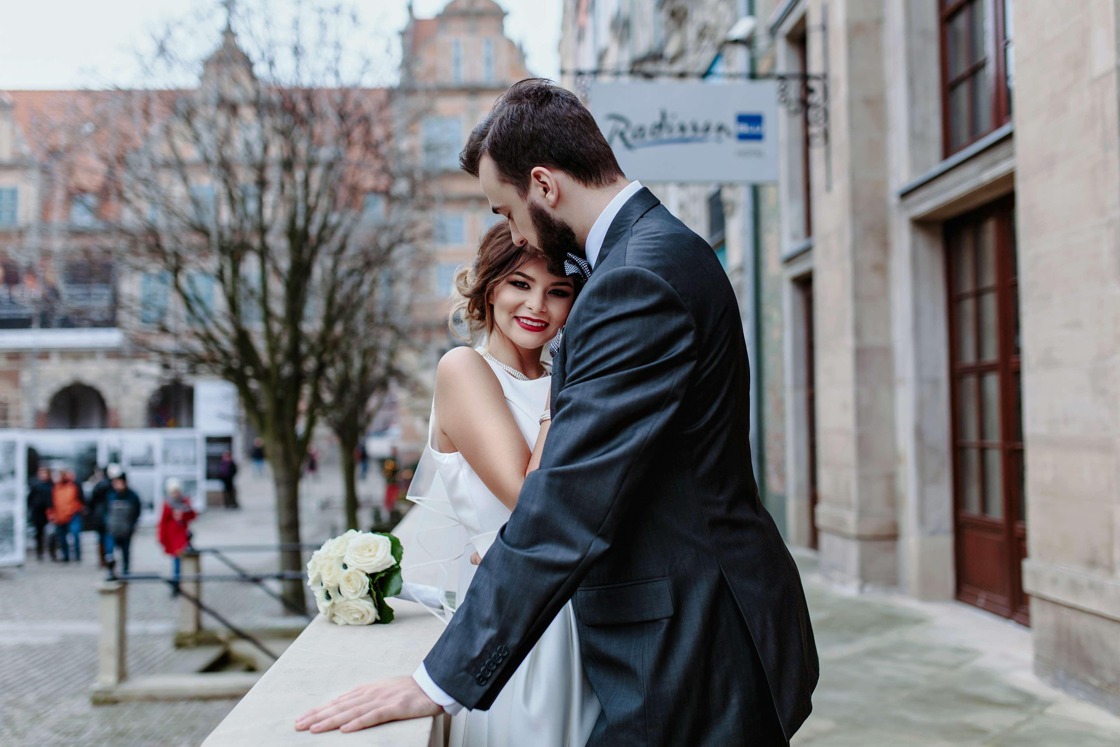 Radisson Blu Hotel, Гданск Екстериор снимка The photo shows a couple embracing in an outdoor setting. The woman, dressed in a white wedding gown, is smiling and appears to be enjoying the moment. The man, wearing a dark suit, leans in toward her, suggesting a romantic atmosphere. In the backgr