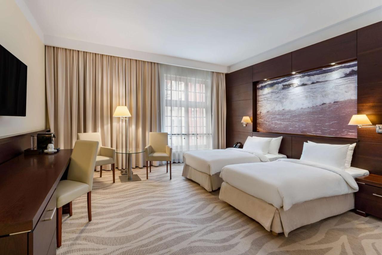 Radisson Blu Hotel, Гданск Екстериор снимка The photo shows a neatly arranged hotel room featuring two beds with white bedding. There is a large window covered with light curtains, allowing natural light to enter the space. In one corner, there are two chairs and a small table, creating a cozy