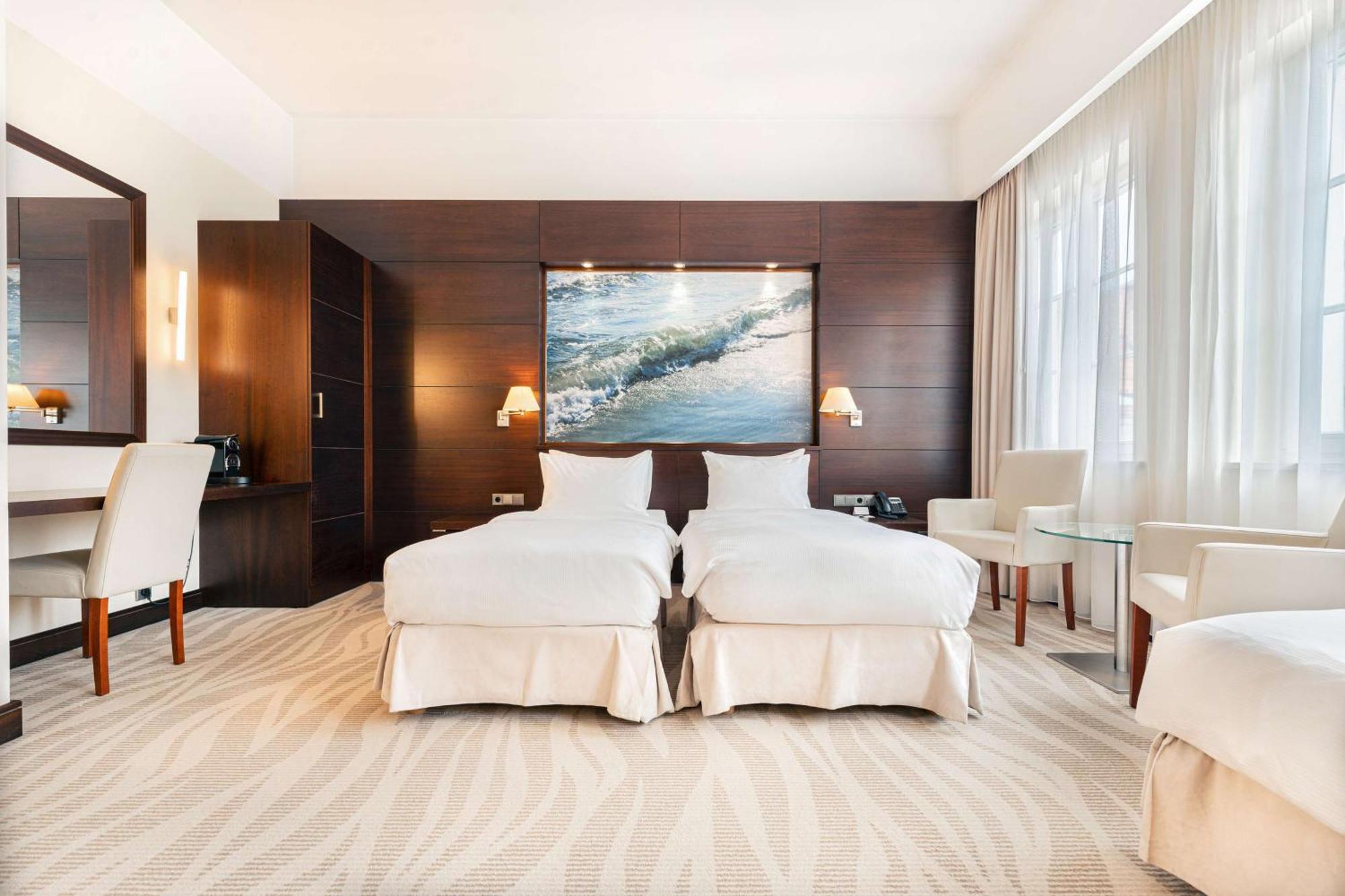 Radisson Blu Hotel, Гданск Екстериор снимка The photo shows a modern hotel room featuring two twin beds dressed in white bedding. The room has a warm, elegant design with wooden cabinetry on one wall and a large piece of artwork depicting a landscape or seascape above the beds. There are two b