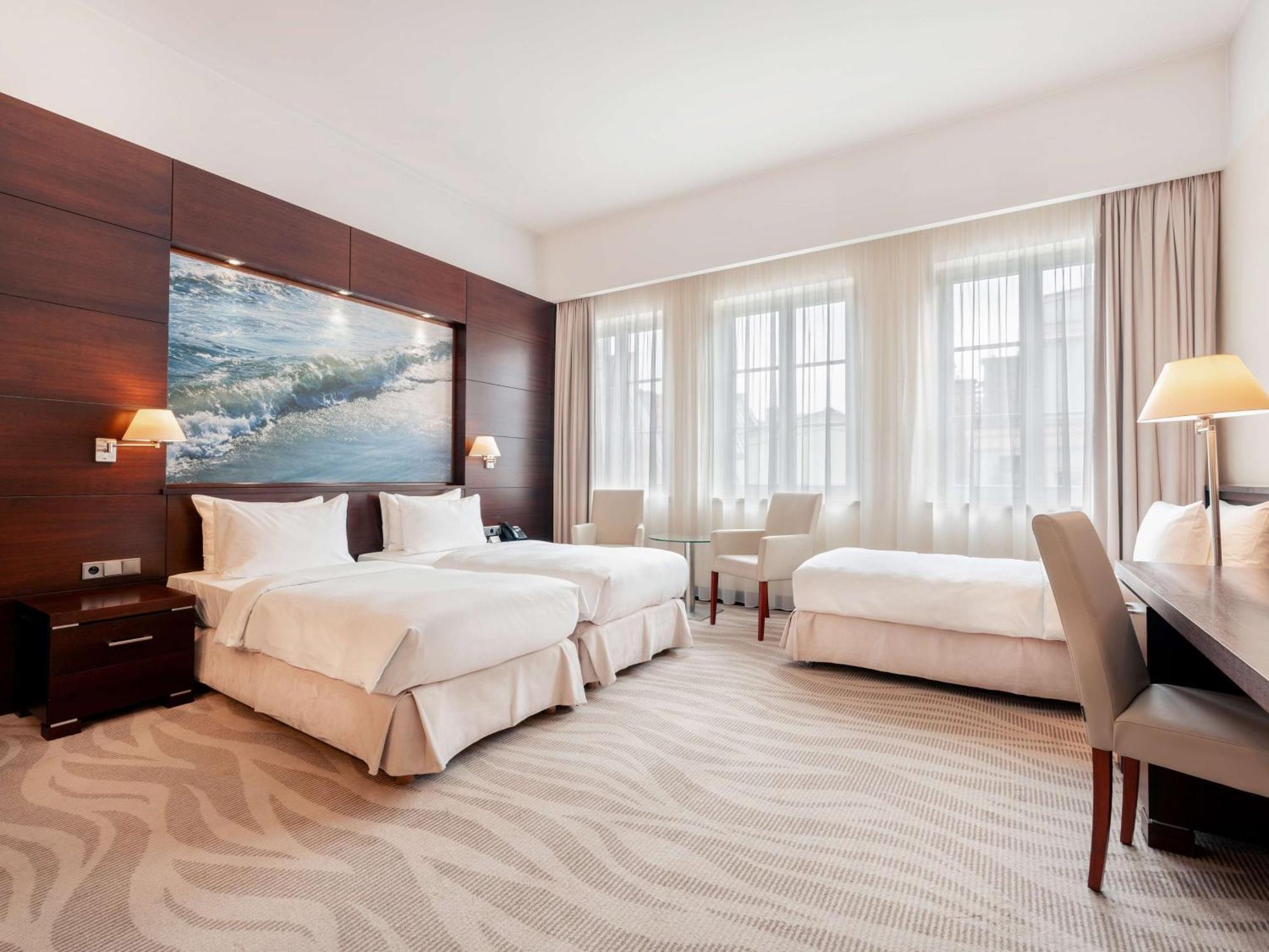 Radisson Blu Hotel, Гданск Екстериор снимка The photo depicts a modern hotel room featuring two twin beds with white bedding, arranged against a wooden wall that includes a large artwork of ocean waves. The room is brightened by natural light streaming through three large windows, which are ad